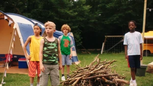 Vier jongens op een camping, tussen hen in ligt een stapel takken en op de achterrond staat een tent.
