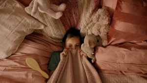 Een foto van en meisje in bed, veel roze dingen, knuffels en kussens, ze trekt een roze deken overzich heen tot aan haar neus.