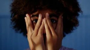 Een close-up foto van een jongen met zijn handen voor zijn ogen, hij kijkt tussen zijn vingers door.