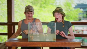 Presentatoren Jurre en Emma kijken vol afschuw naar een bak water die voor hen op tafel staat, er zitten kwallen in.