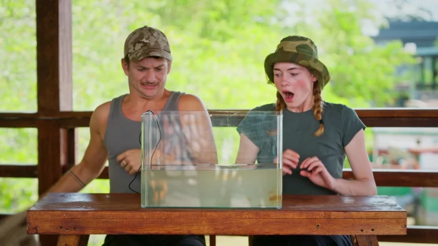 Presentatoren Jurre en Emma kijken vol afschuw naar een bak water die voor hen op tafel staat, er zitten kwallen in.