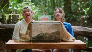 Een foto van de beide presentatoren, voor het staat een grote glazen bak waarover een doek zit. Jurre moet lachen, Emma heeft een soort verband/touw om haar neus en kin en hoofd.
