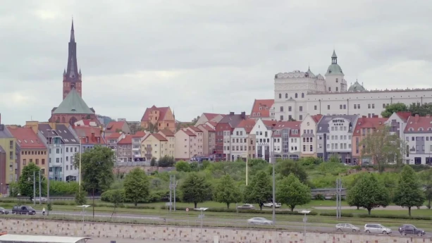 Een foto van een stadbeeld, je ziet een kade met daarachter een kerk, geel en wit gekleurde huizen met rode daken en een paar waar auto's voor langs rijden.