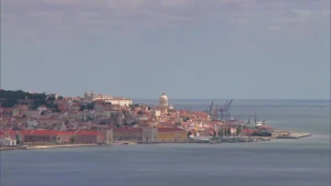 Een foto van een stadje aan de kust, de witte kerktoren licht op in de zon, het stadje steekt een stukje de zee in, je ziet huizen, kerken, kranen en linksachter een park of bos.