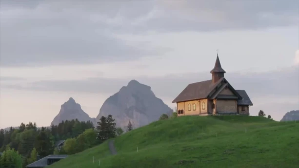 Een foto van een Zwitsers landschap met een kerkje op een groene heuveltop, daarachter hoge grijze bergen.  