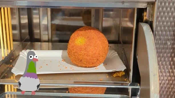 Een foto van een eierbal, een krokante ronde snack. Hij ligt in een vakje bij de snackbar waar je hem 'uit de muur' kan trekken. Linksonder staat een getekende duif.