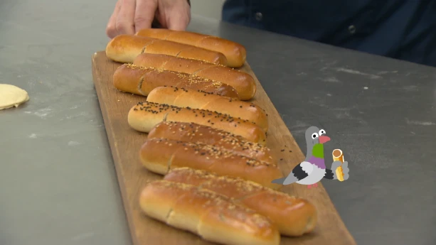 Een foto van gesneden worstenbroodjes op een houten plank. Op de plank zit een getekende duif, die een klein worstenbroodje eet.