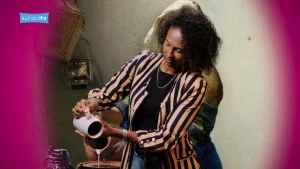 De presentatrice doet het melkmeisje na, achter haar is het schilderij te zien