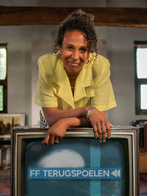 Presentatrice Astrid Sy staat op een oude, bruine zolder. Ze leunt over een televisie waar 'FF terugspoelen' in beeld staat.