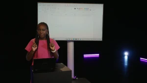 Een foto van presentatrice Tirsa die achter een laptop staat. Achter haar staat een groot scherm. Ze steekt haar vingers op ten teken dat je moet opletten op wat ze gaat typen. Er staan nog geen woorden op het scherm.