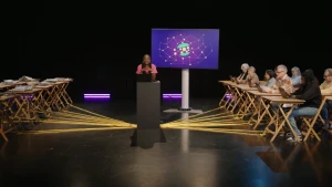 Een foto van presentatrice Tirsa die middenin een zaal achter een laptop staat. Links van haar tafels met boeken en kranten erop. Rechts van haar tafels met mensen achter laptops. Alle tafels zijn verbonden met waar zij staat door middel van gele touwen.