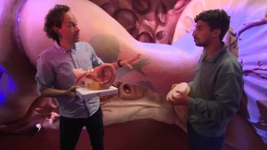 Een foto van presentator Nizar en een oorarts. De oorarts houdt een model van een oor vast. Op de achtergrond is een sterk uitvergroot onderdeel van het oor te zien, het trommelvlies. Nizar houdt een trommeltje vast.