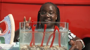 Een foto van presentatrice Tirsa die bij een glazen bak gevuld met water zit. In de bak staan rode draden recht overeind onder water. Links van haar is een stuk van een model van het oor te zien.