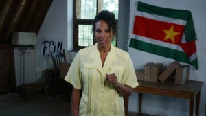 Presentatrice Astrid staat in het midden van het beeld. Achter haar hangt de vlag van Suriname.