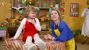 Een foto van de presentatrice en een meisje, het meisje zit op tafel met de gemaakte ketting om haar nek. 