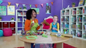 Een foto van de presentatrice en een meisje ze zitten aan tafel te knutselen en moeten erg lachen.