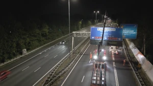 Een foto van een snelweg 's avonds in het donker. Op de snelweg staat een werkauto met een hijskraan. De kraan takelt een blauw bord met daarop plaatsnamen en de afstand omhoog. Op de weg lopen mensen in oranje veiligheidshesjes.
