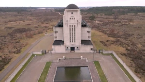 Een foto gemaakt met een drone van een hoog betonnen gebouw met kleine raampjes. het middelste gedeelte van het gebouw heeft een koepelvormig dak. Voor het gebouw ligt een vierkante vijver. Om het gebouw heen is alleen maar heide te zien.