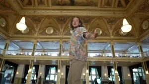 Een foto van presentator Pascal die in de hal van een hotel staat. Het dak is rijk versierd met schilderingen en er hangen grote luxe lampen: kroonluchters. Pascal heeft zijn camera uitgestrekt naar de camera alsof hij je uitnodigt mee te gaan.