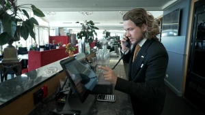 Een foto van presentator Pascal in een net zwart pak. Hij staat achter een bar in een hotel en heeft een telefoon aan zijn oor om een roomservice-bestelling op te nemen. Hij klikt iets aan op een beeldscherm.