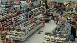 Een foto van een supermarkt van bovenaf gezien, je ziet verschillende koelvakken en een paar mensen die bij de schappen staan.