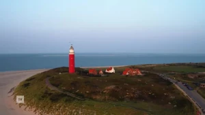 Een foto vanuit vogelperspectief van de vuurtoren van Texel, je zie de zee, het strand en het deel van het eiland met de vuurtoren en enkele huizen.