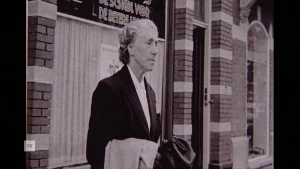 Een zwart-wit foto van gebedsgenezeres Greet Hofmans, ze staat buiten voor een gebouw met een jas over haar arm.