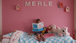 Meisje zit op een bed en leest een boek. Achter haar een roze muur met haar naam, 'Merle' erop.