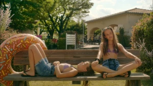 Twee meiden zitten op een bankje. Het is zomer. De een ligt en de ander zit in kleermakerszit.