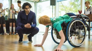 Jongen met groen shirt in roelstoel komt met zijn handen bij de grond. Een andere man zit gehurkt en spreekt met hem.