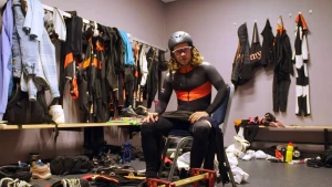 Een foto van presentator Pascal die in een sportkleedkamer zit. Hij draagt een strak schaatspak, een helm en een beschermende bril. Op de grond liggen schaatsen.