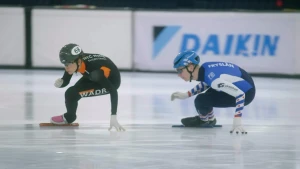 Een foto van twee schaatsers die snel door de bocht gaan op een schaatsbaan. Ze leunen beide in de bocht en houden hun hand op het ijs.