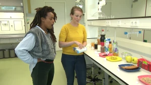 Presentatrice Eva en een vrouw staan in een laboratorium. De vrouw heeft een doos suikerklontjes in haar handen. Ze kijken naar een tafel met borden en glazen met verschillende soorten eten en drinken, zoals ontbijtkoek, druiven en chocolademelk.