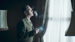 Een foto van een man in groen uniform, hij staat in een donkere kamer voor een raam een sigaret te roken.