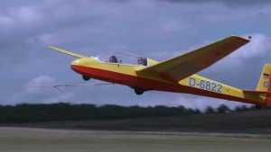 Een foto van een zweefvliegtuig, een geel en rood vliegtuig zonder motoren. Er zitten twee mensen in. Het vliegtuig wordt de lucht in getrokken door een kabel.