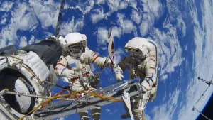Foto van twee astronauten die buiten het ruimtestation aan het werk zijn, de achtergrond zie je de blauwe aarde met witte wolken.
