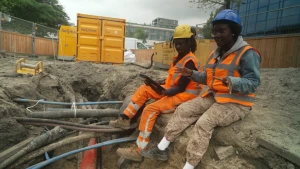 Een foto van presentatrice Tirsa en een kabelwerker. Ze zitten op de rand van een gat in de grond waar allemaal kabels en leidingen lopen. Ze dragen een bouwhelm en oranje reflecterende kleding.