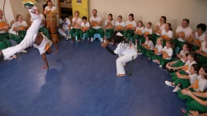 Een foto van twee dansende mannen in een zaaltje. Eén van de mannen staat op zijn handen. Om hen heen een groep mensen in een cirkel, ze dragen allemaal een groene broek en een wit shirt met logo. Sommige mensen klappen, anderen spelen trommel.