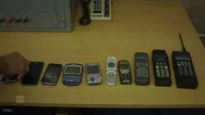 Een foto van een tafel waarom 9 oude mobiele telefoons liggen, links de grootste, rechts de kleinste, een hand legt er een tiende telefoon bij.