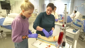 Janouk staat in het ziekenhuis met een studente naast zich. Ze hebben handschoentjes aan en oefenen op een kunstarm met bloedprikken.
