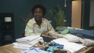 Een foto van presentatrice Tirsa die aan een tafel zit, daarop liggen allerlei kledingstukken en andere dingen van katoen.