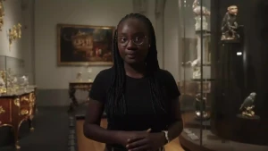 Thirsa staat in een ruimte met schilderijen en vitrines om zich heen. Er zijn veel gouden details achter haar te zien.