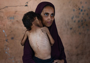 Foto van gesluierde vrouw met een mager kind op haar arm, gezien op de rug. 