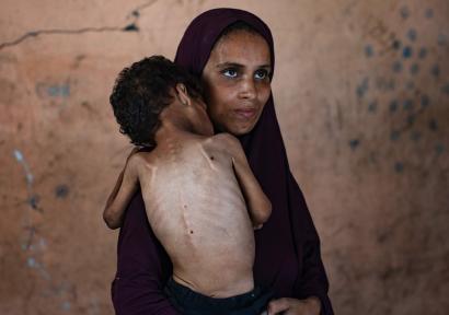 Foto van gesluierde vrouw met een mager kind op haar arm, gezien op de rug. 
