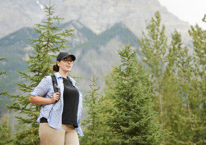 Foto van vrouw met rugzak en pet in een berglandschap met groene bomen op de achtergrond. 