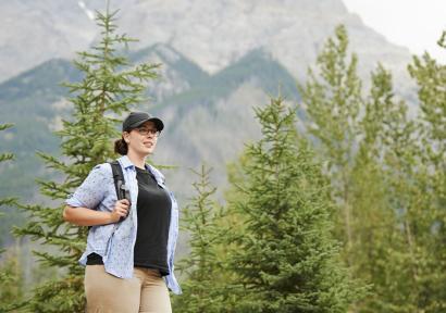 Foto van vrouw met rugzak en pet in een berglandschap met groene bomen op de achtergrond. 