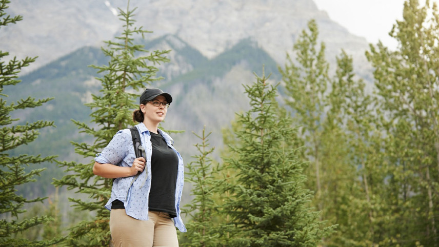 Foto van vrouw met rugzak en pet in een berglandschap met groene bomen op de achtergrond. 