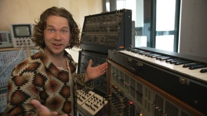 Een foto van presentator Pascal in een muziekstudio. Voor hem staat een keyboard, om hem heen staan diverse apparaten met allerlei knopjes.
