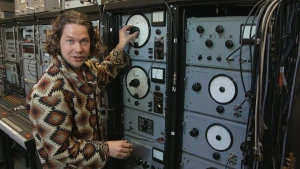 Een foto van presentator Pascal bij radio-apparatuur uit de jaren zestig. Op de apparatuur zitten knoppen en metertjes die waardes aangeven. Pascal draait aan een knop.