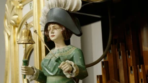Foto van mannetje met hoed en bel op draaiorgel, houten buizen en goudkleurige versiering zijn zichtbaar op de achtergrond.